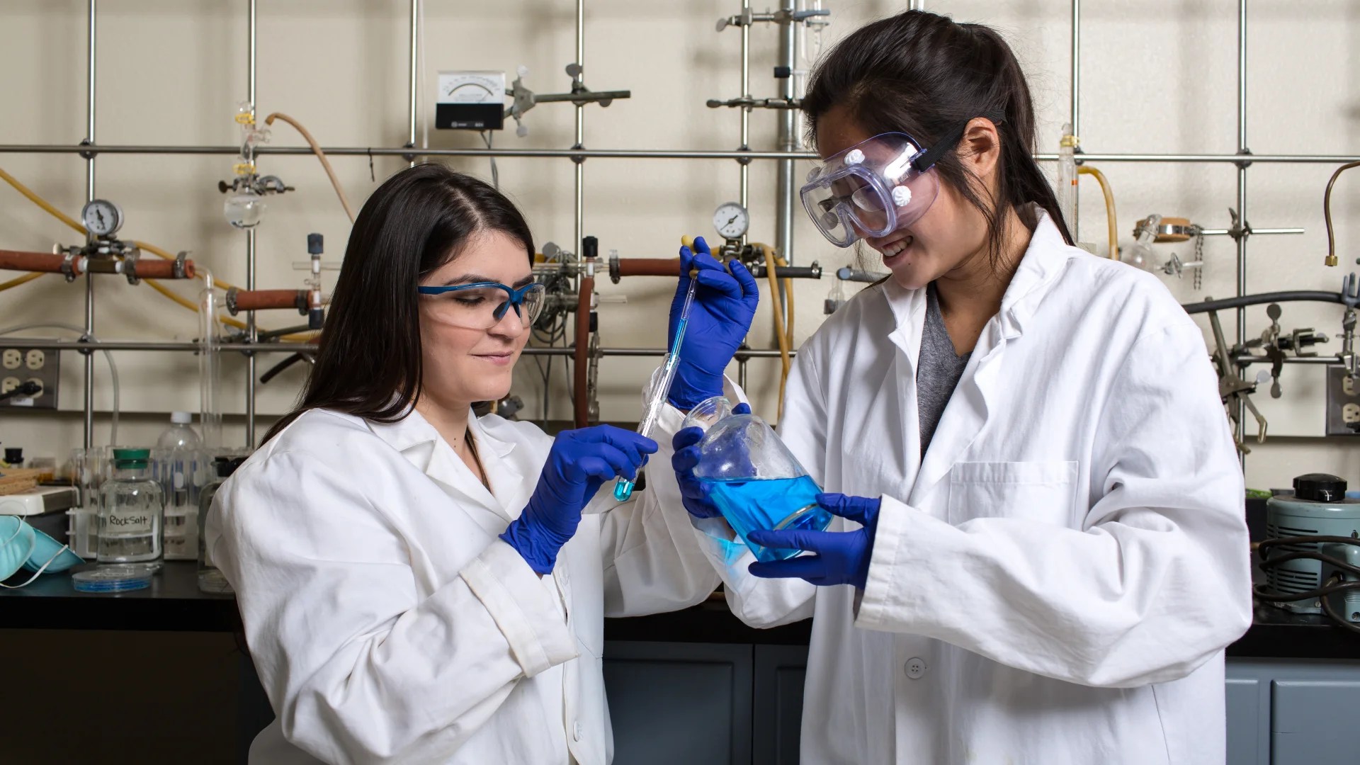 Two people wearing lab coats, gloves, and goggles working together to mix a blue chemical solution in a laboratory