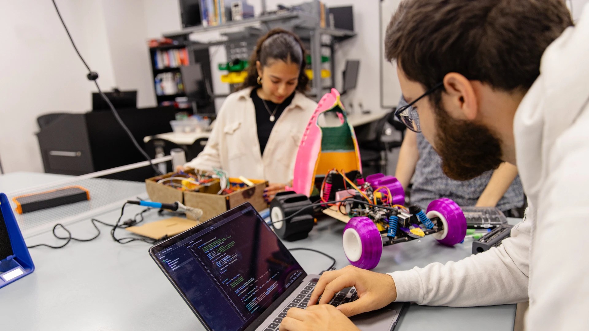 People working together on a robotics project, with one person programming code on a laptop and others assembling a robotic car