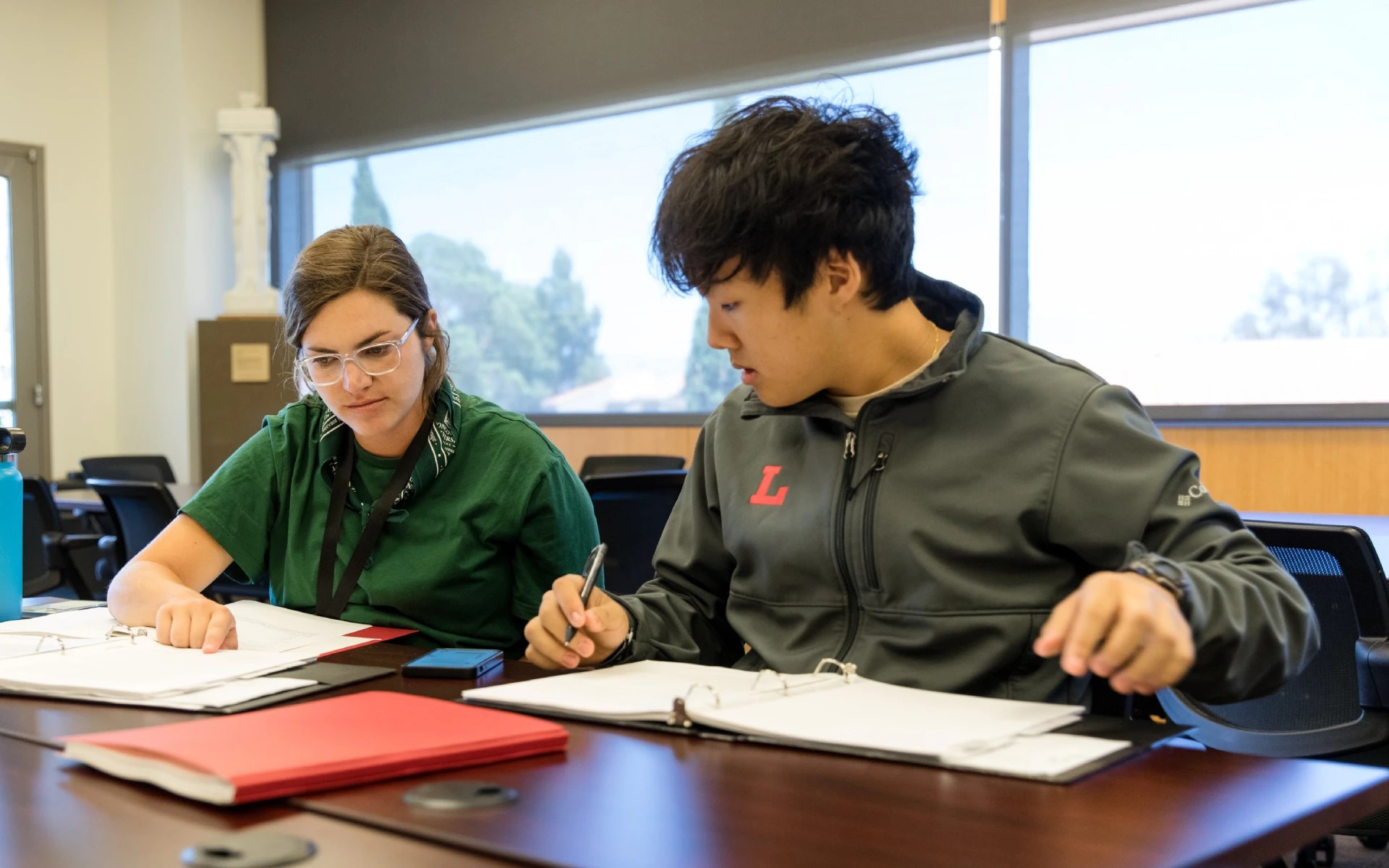 Two people sitting at a table looking at binders and taking notes during a study session