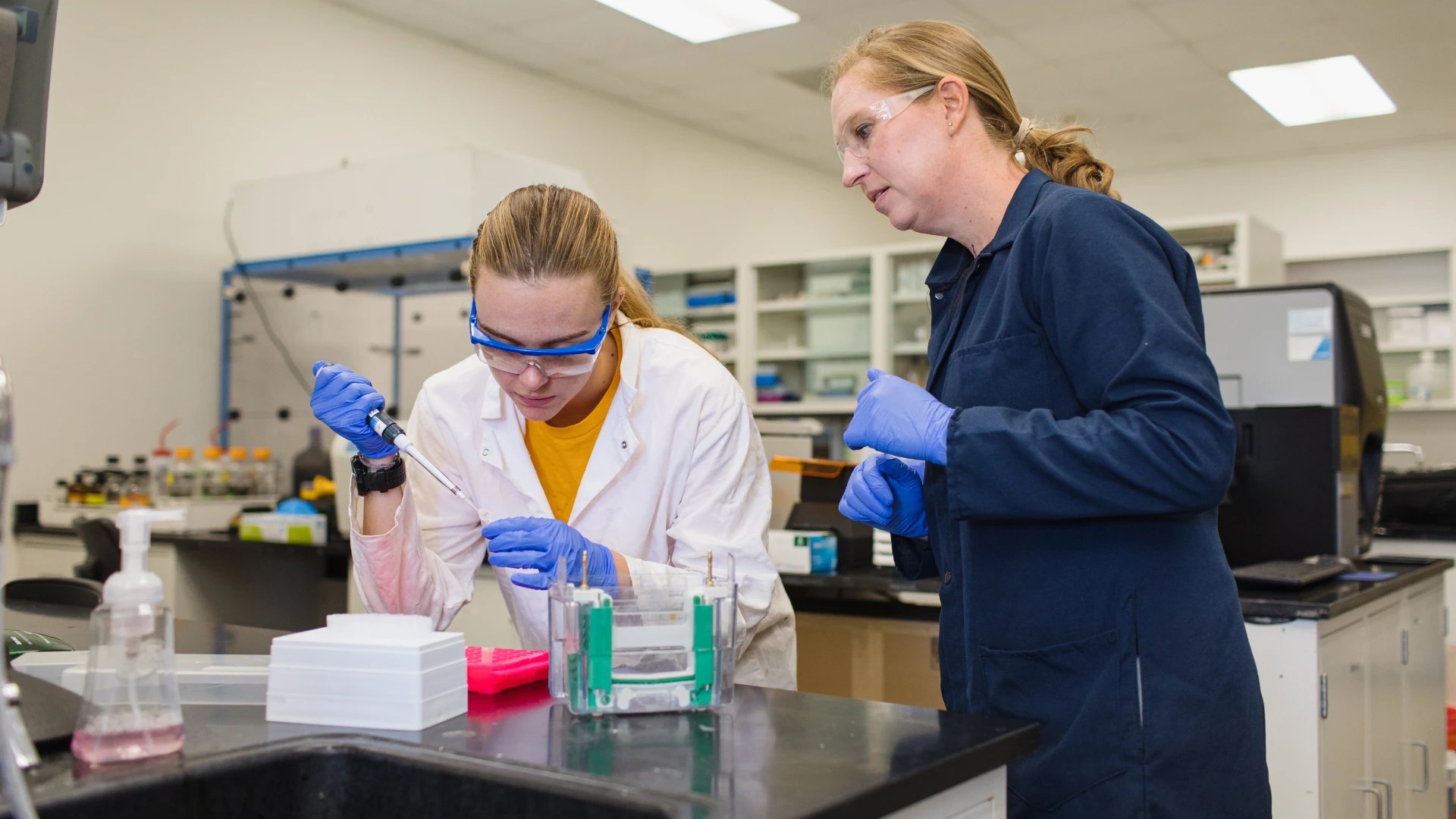 Two people in lab coats and gloves using lab equipment and pipettes on a black countertop
