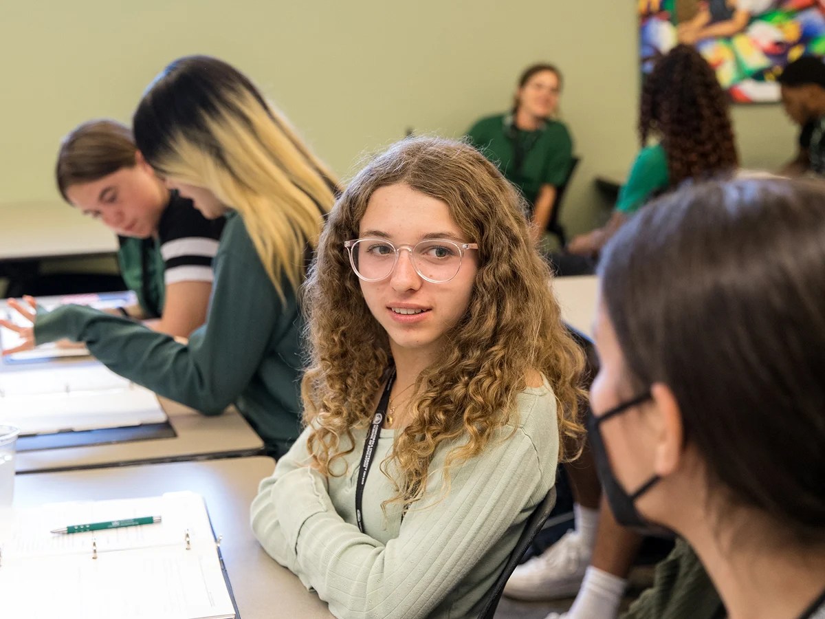 A girl sitting at a desk with other people in the background