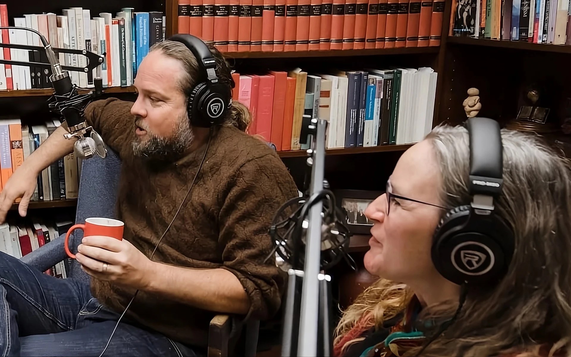 A man and woman sitting in chairs with microphones and bookshelves