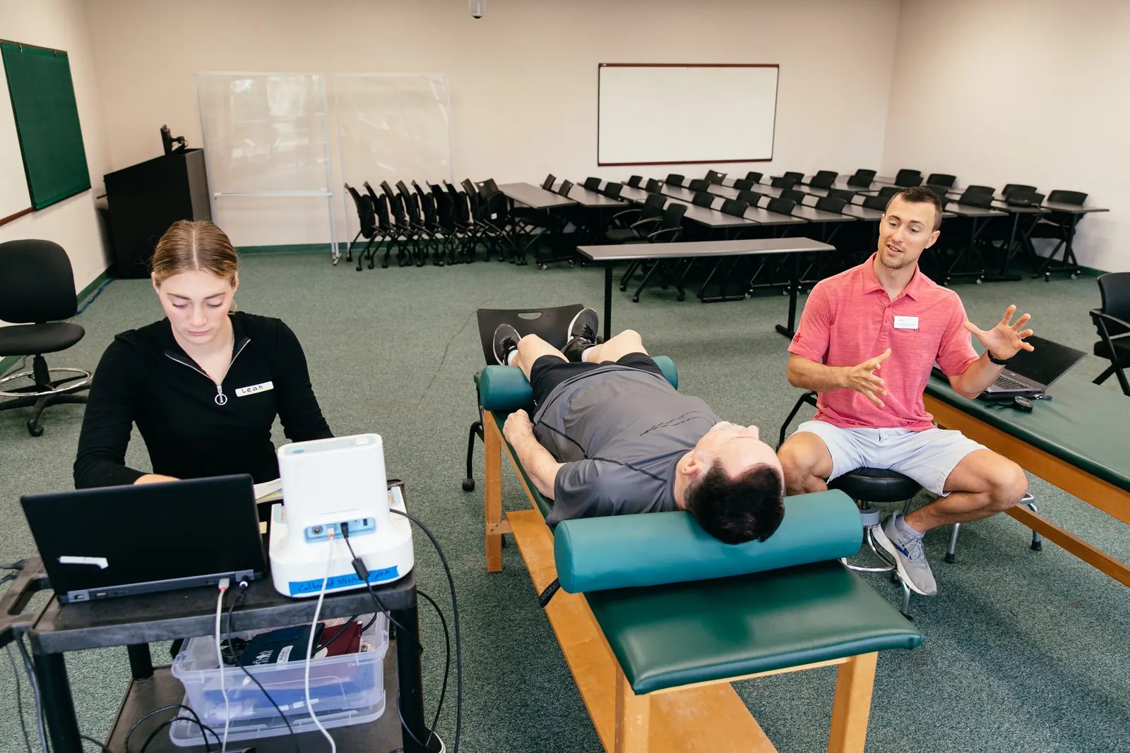 A student research assistant records data on a laptop while a researcher talks with a participant lying on an exam table.