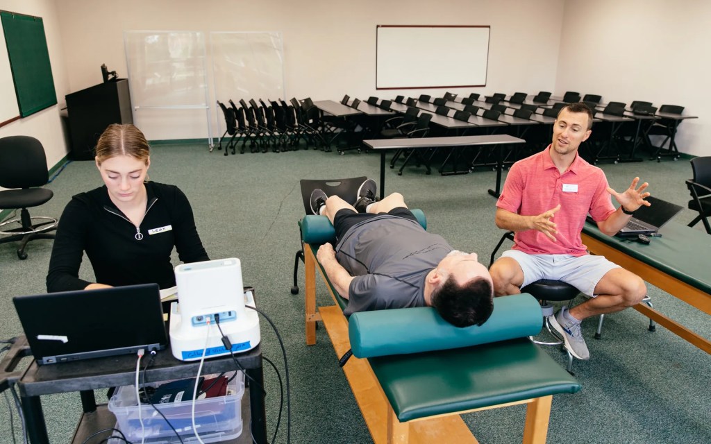 A student research assistant records data on a laptop while a researcher talks with a participant lying on an exam table.