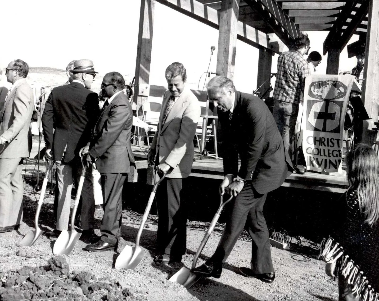 Black and white photo of men in suits breaking ground with shovels beside the Bird Sanctuary stage and a Christ College Irvine banner.