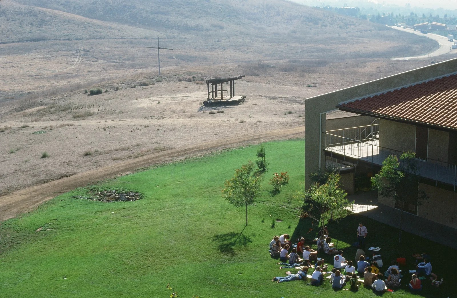 The Bird Sanctuary and a tall cross on an open hillside next to an early campus building, with a group seated on the lawn below.